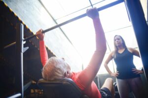 Senior man bench pressing with a female trainer overseeing his workout in an indoor gym.