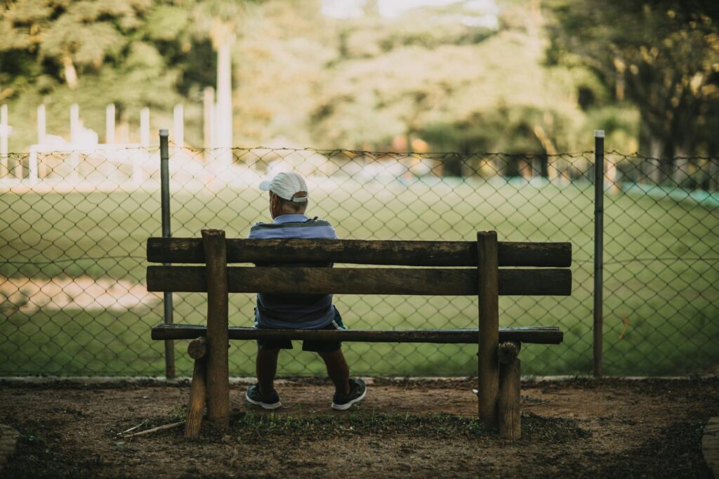 A senior man in a white cap sits alone on a wooden bench, enjoying the peaceful surroundings of a park.