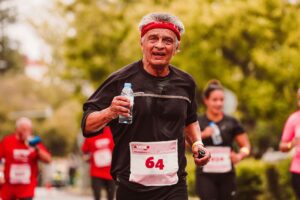 Elderly man running a marathon outdoors, holding a water bottle and smiling.