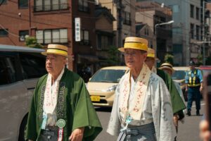 Elderly men in traditional attire participate in a cultural parade during a sunny day in Tokyo.