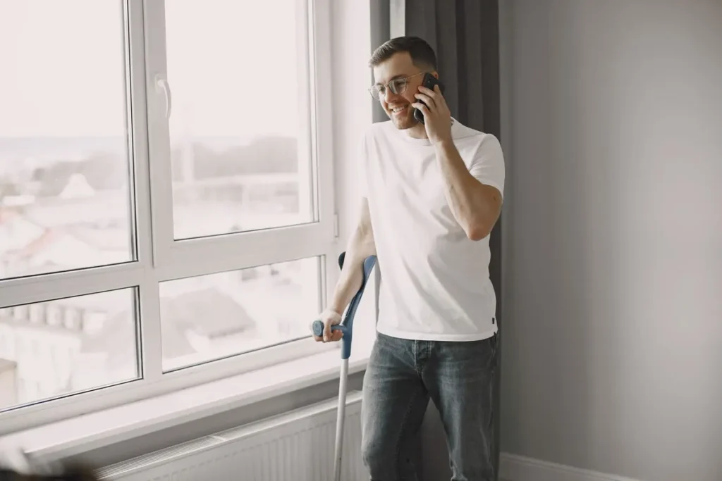 Young man standing by the window on crutches, talking on smartphone with a smile.