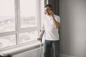 Young man standing by the window on crutches, talking on smartphone with a smile.