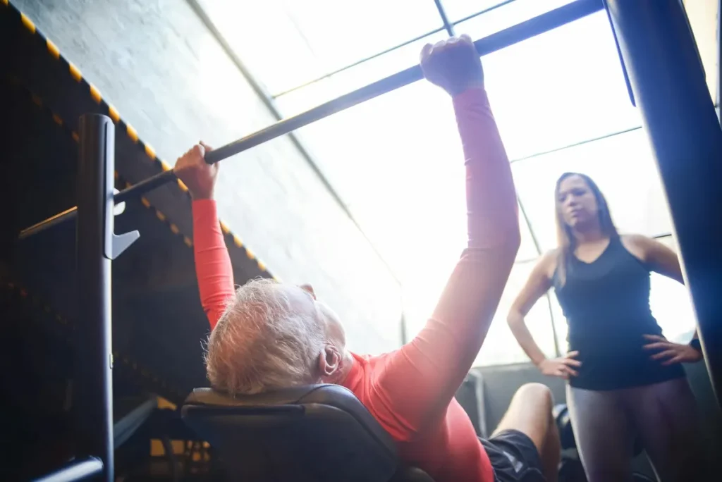 Senior man bench pressing with a female trainer overseeing his workout in an indoor gym.