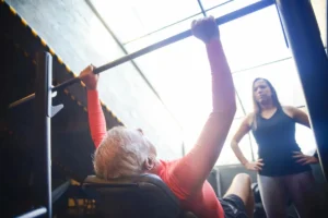 Senior man bench pressing with a female trainer overseeing his workout in an indoor gym.