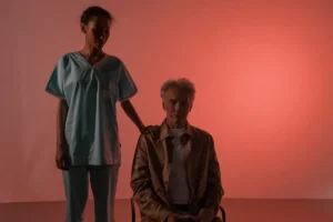 A nurse standing beside an elderly man in a studio with a warm background.