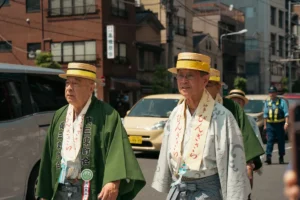 Elderly men in traditional attire participate in a cultural parade during a sunny day in Tokyo.