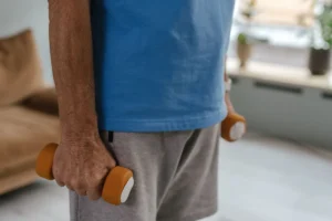 A senior holding orange dumbbells, indoors in casual exercise attire.