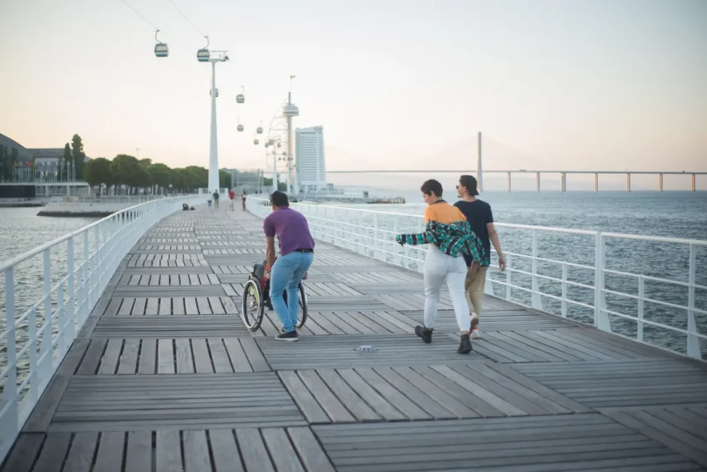 Three friends enjoy a sunset walk along a scenic Lisbon boardwalk with a wheelchair.